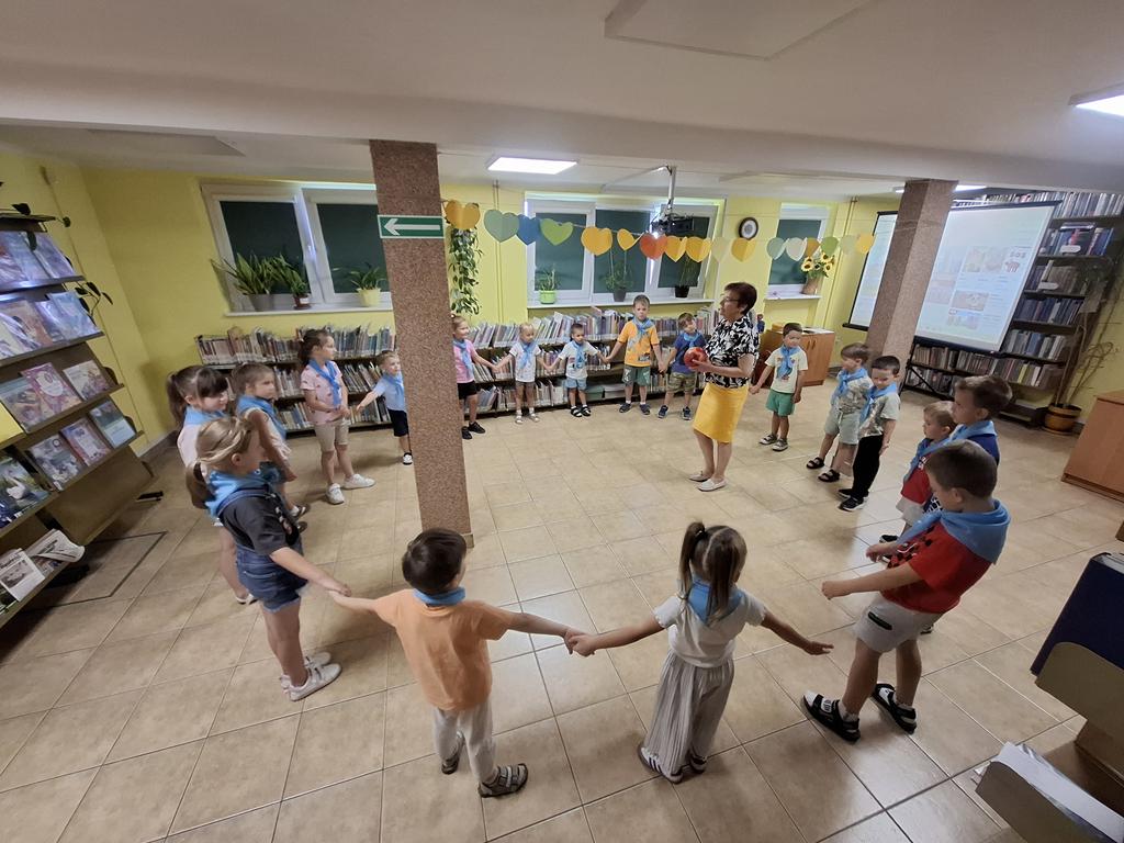 Grupa przedszkolaków w kolorowych ubraniach i niebieskich apaszkach stoi w okręgu. Dzieci trzymają się za ręce. Na środku kręgu stoi bibliotekarka w żółtej spódnicy i czarno-białej bluzce trzymająca w ręku piłkę. W tle są regały z książkami.
