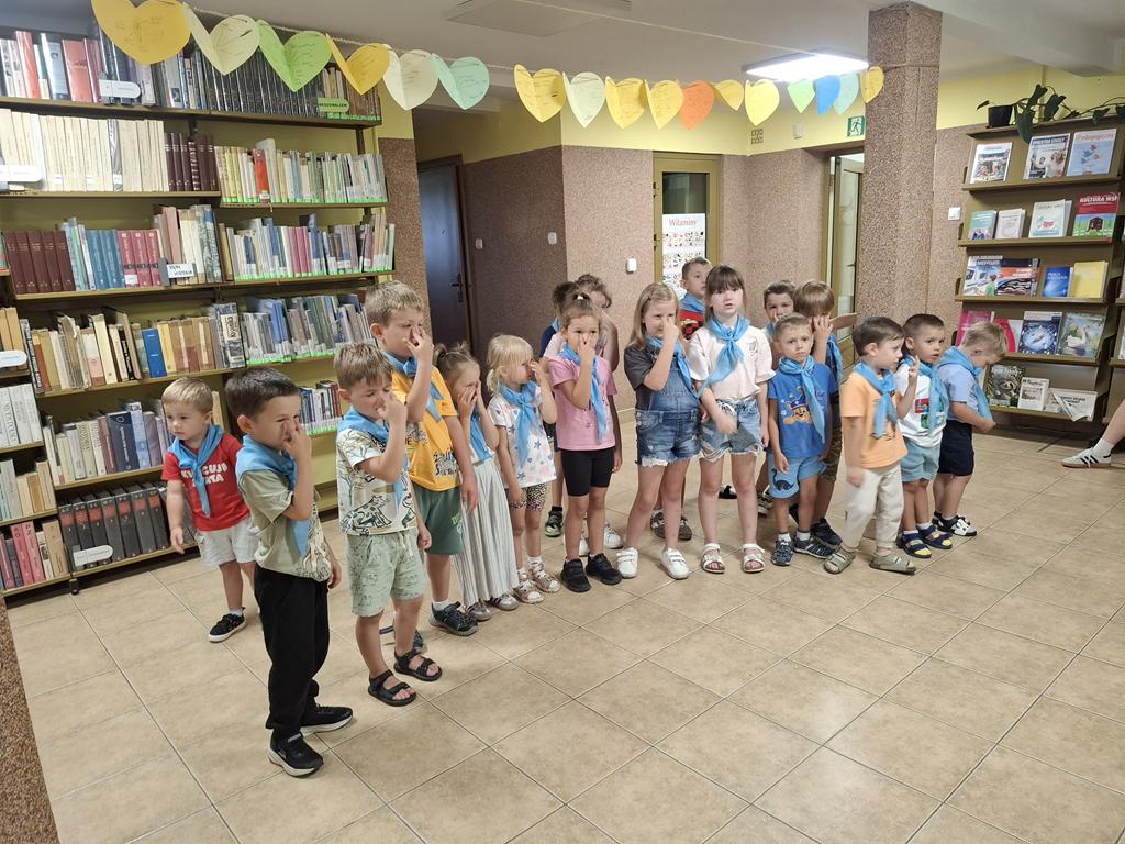 Grupa przedszkolaków w kolorowych ubraniach i niebieskich apaszkach stoi w rzędzie i trzyma się za nos. W tle są regały z książkami.
