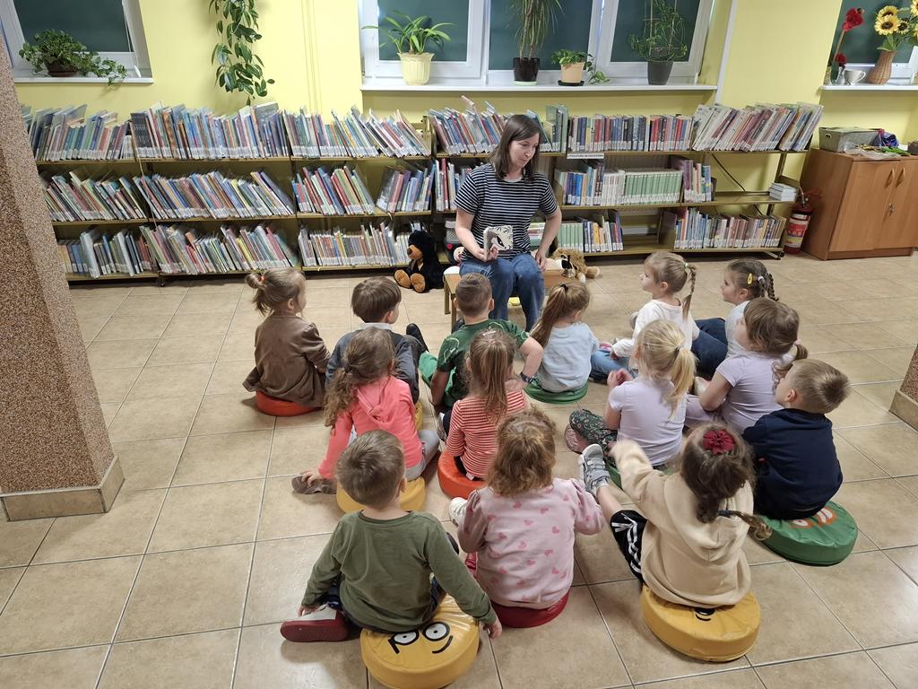 Kobieta w bluzce w czarno białe paski i niebieski spodniach siedzi na ławeczce. W ręku trzyma książkę "Rok w lesie. Borsuk". Przed kobietą siedzi grupa dzieci w kolorowych ubraniach. Za kobieta są regały z książkami i leżą na podłodze maskotki.
