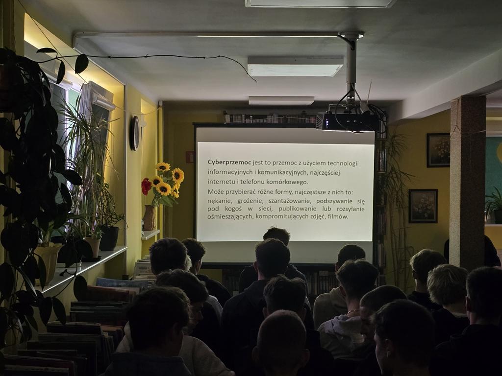 Na ekranie rzutnika jest opisane co to jest cyberprzemoc. Przed ekranem siedzi grupa młodzieży.