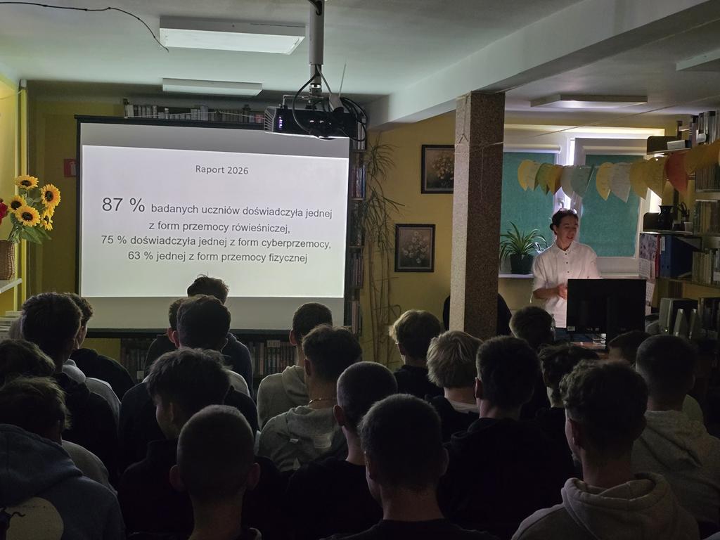 Na ekranie rzutnika jest wyświetlony raport z 2026 roku dotyczący przemocy. Po prawej stronie od ekranu stoi kobieta w białej bluzce. Przed ekranem siedzi grupa młodzieży.
